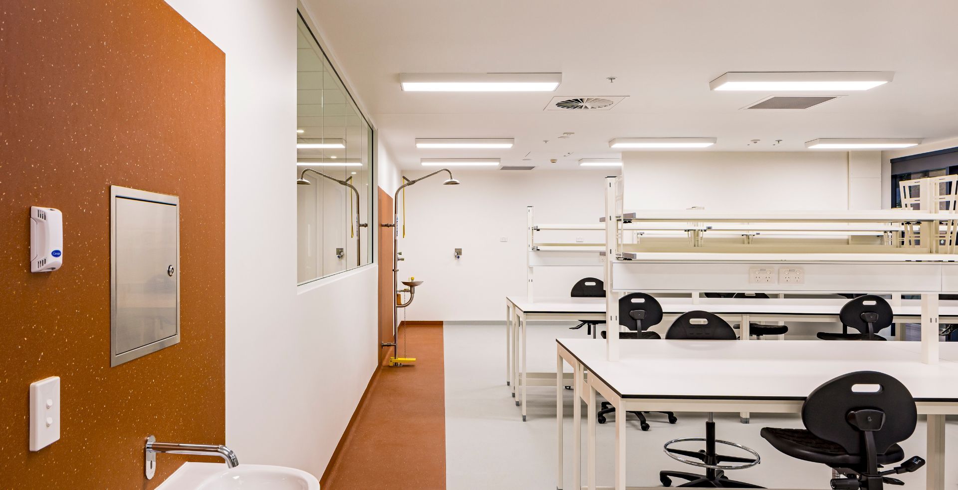 Orange feature wall in a PC2 laboratory, fitted with a safety shower and eyewash, opening onto a row of lab benches and stools