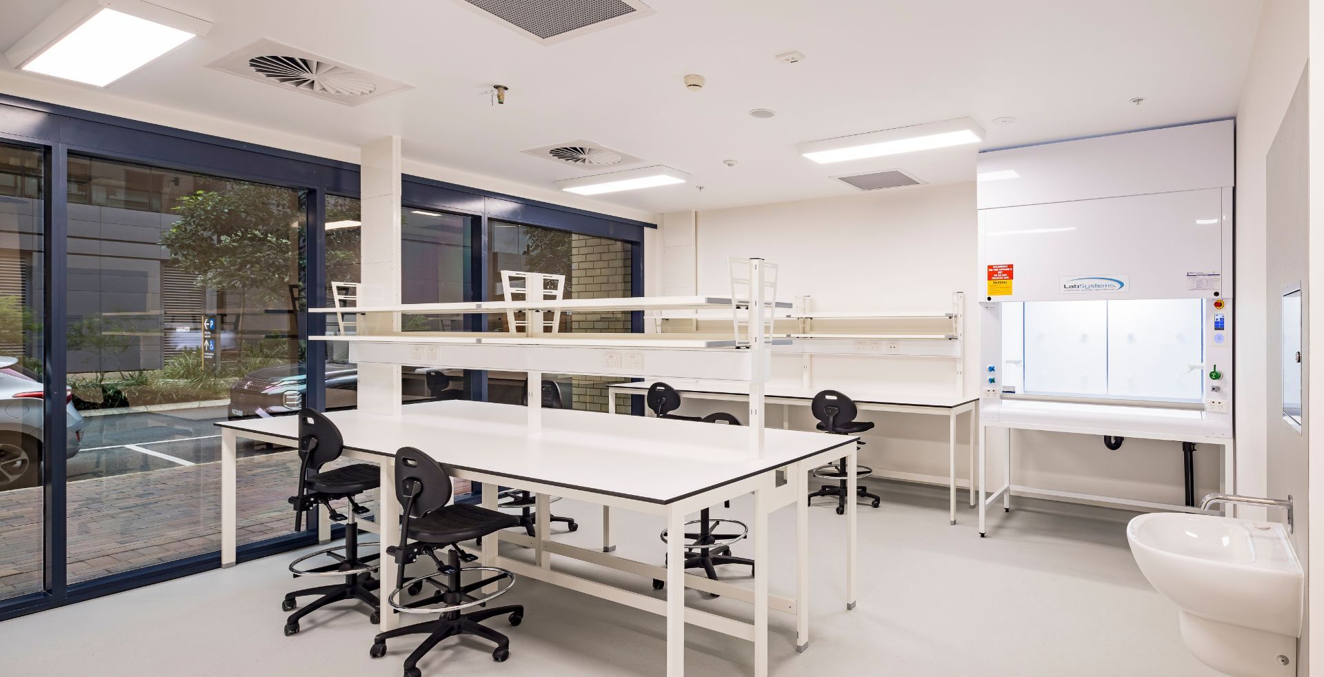 Interior of a PC2 laboratory showing a fume cupboard, sink and open bench seating beneath large street-facing windows