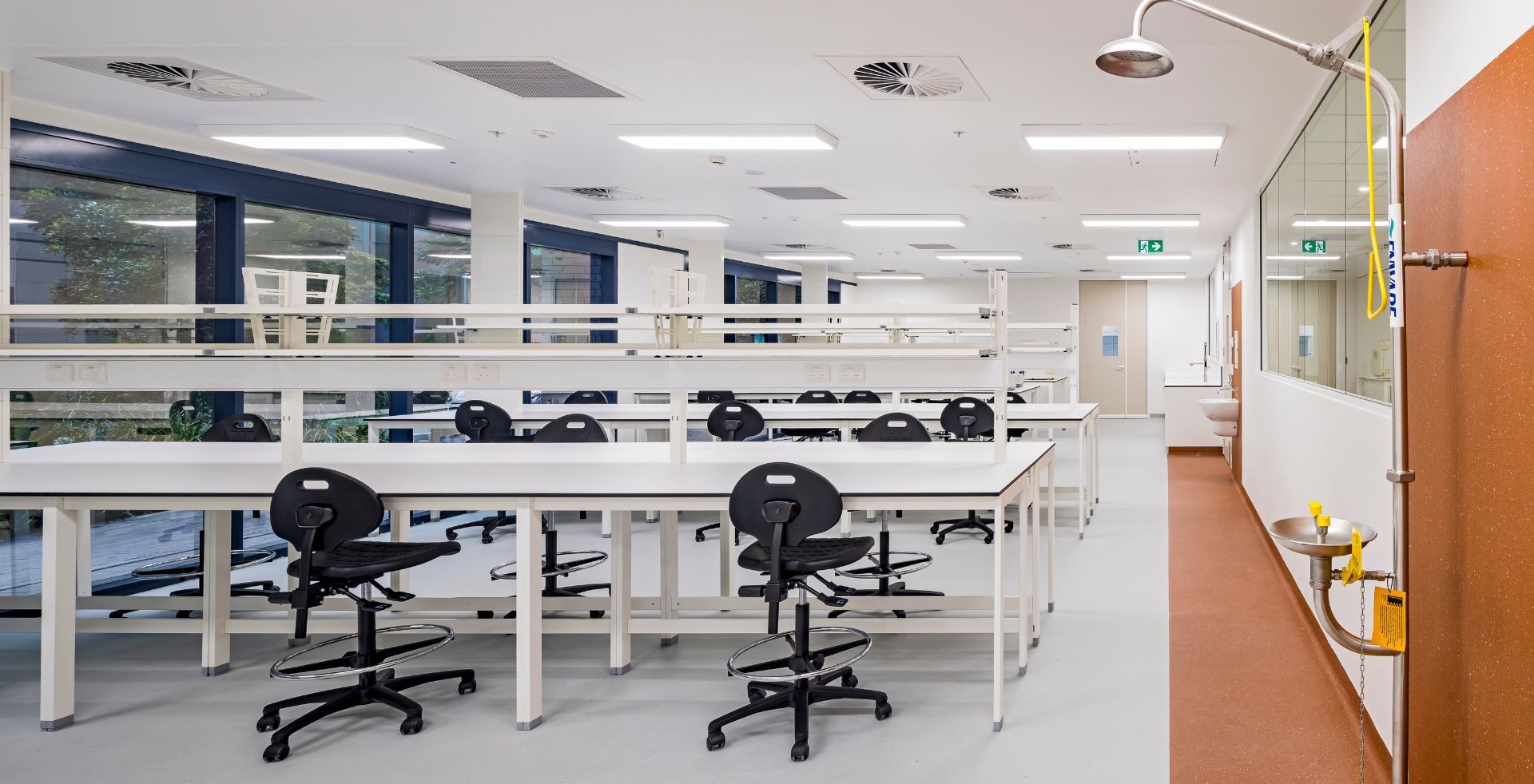 Rows of laboratory benches and stools in a PC2 lab, leading to a safety shower on an orange accent wall