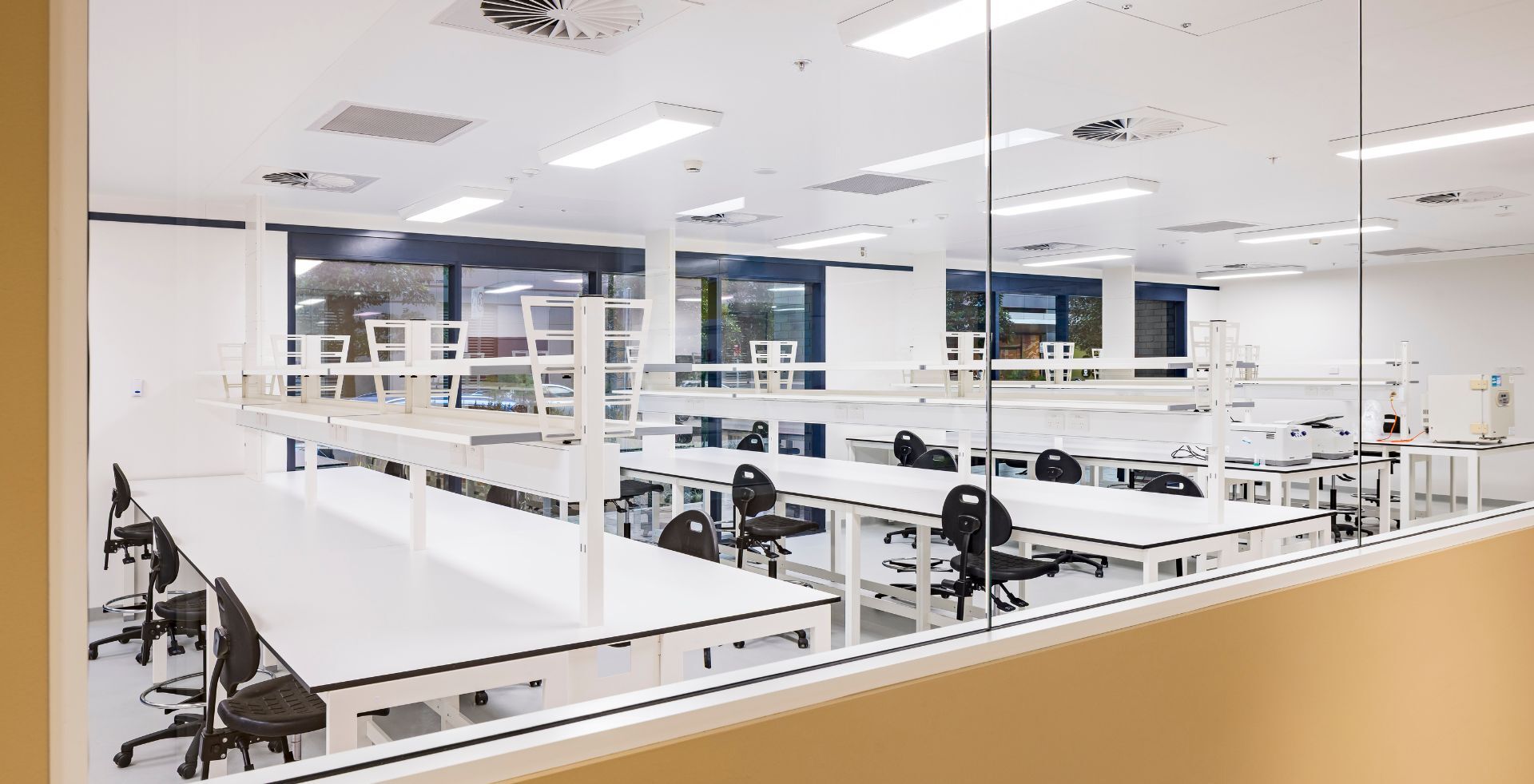 View through a glass partition into a PC2 laboratory fitted with benches, stools and lab equipment, with windows along the far wall