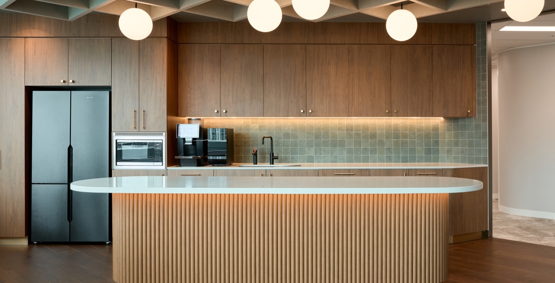 Office kitchen and breakout area featuring a long timber-batten island bench, matching wall cabinets, and integrated appliances.