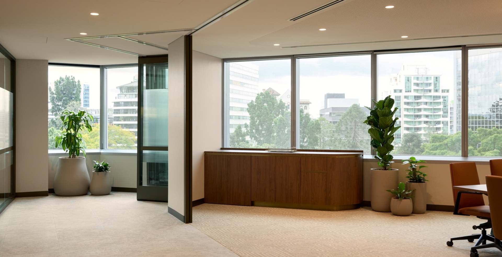 Breakout space with large open view windows framed by large potted indoor plants and a curved timber credenza.