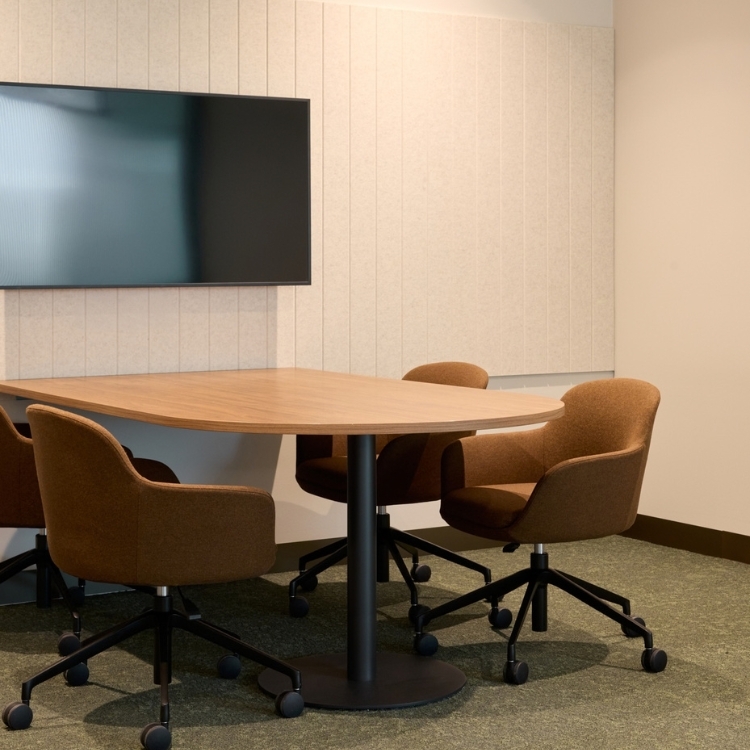 Small collaborative meeting space with a timber table, brown fabric swivel chairs, and a wall-mounted display screen.