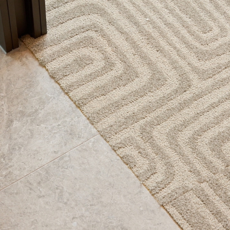 Textured beige carpet with a repeating geometric pattern meeting a polished light gray tile floor border.