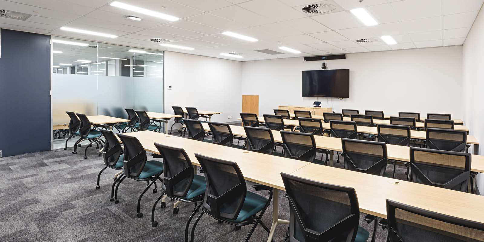 Lecture theatre with many rows of tables and chairs with a TV