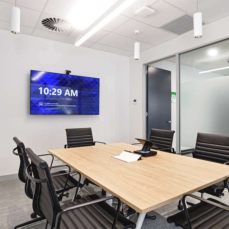 A modern conference room with a wooden table surrounded by six black chairs on wheels, reminiscent of a sophisticated Bio-Rad laboratory. A television mounted on the wall displays 