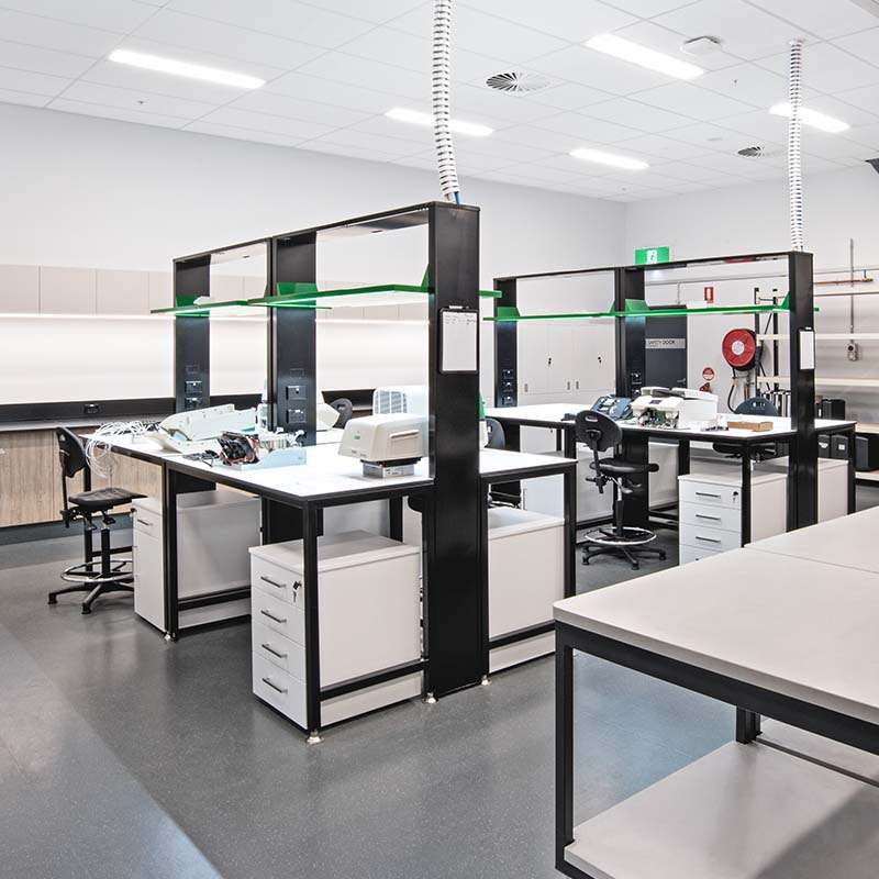A modern, well-lit laboratory with multiple workstations equipped with chairs, cabinets, and various scientific instruments. The workspace features black and white furniture, green overhead lighting fixtures, and a gray floor reminiscent of an organized warehouse. Emergency equipment is visible in the background.