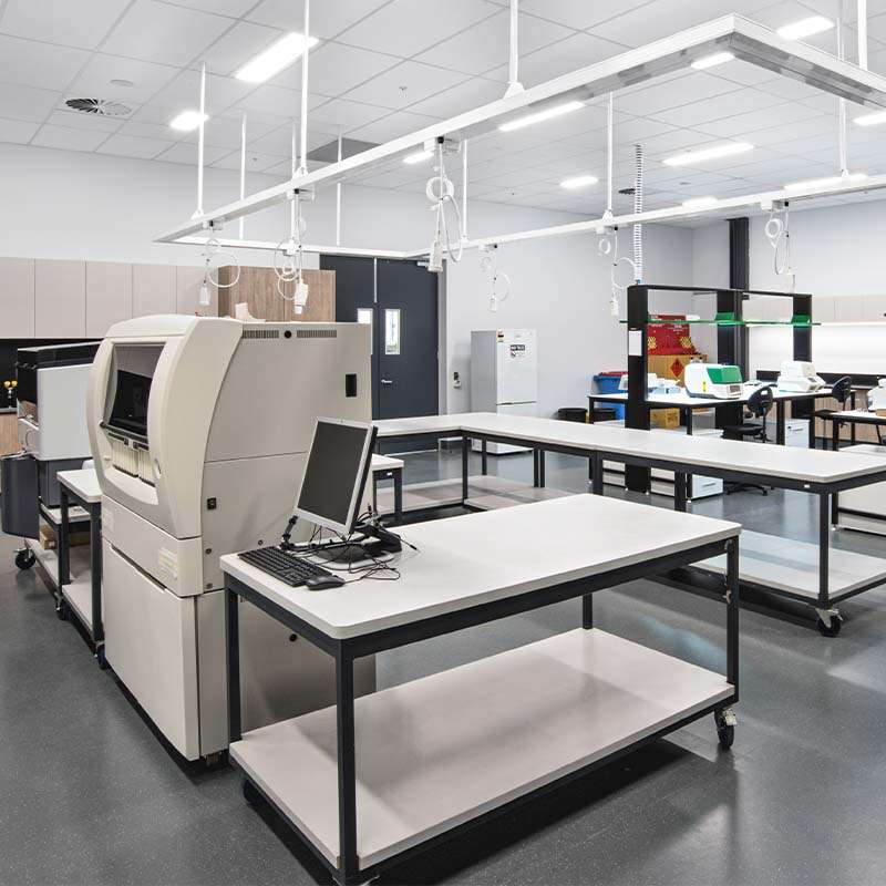 A modern laboratory with several workstations equipped with computers, various lab equipment, and machines. The room has bright lighting, white walls, and a clean, organized setup reminiscent of a well-structured distribution centre. Several hanging power outlets are visible.