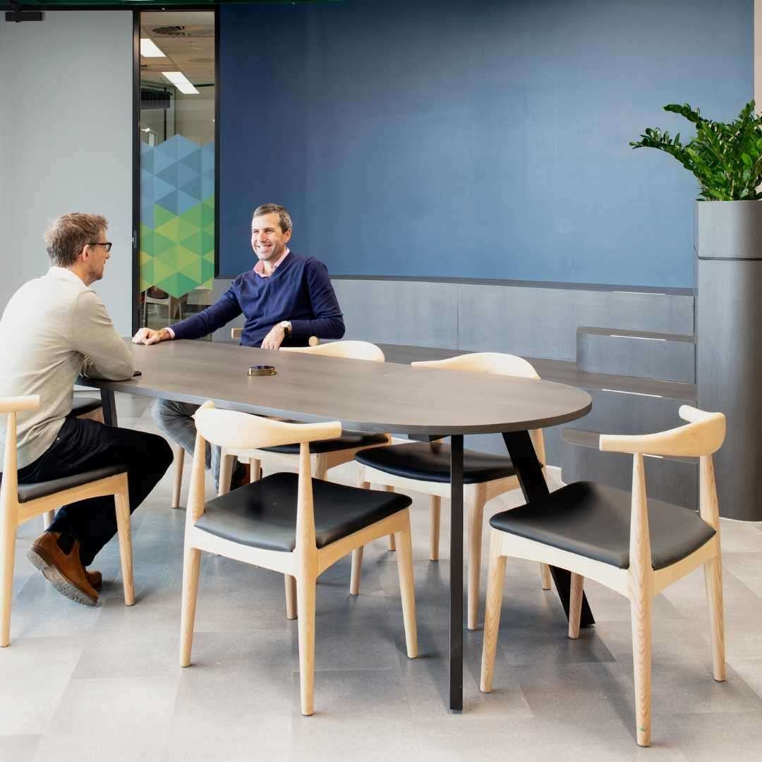 A person leans on a balcony railing on a modern patio, overlooking the city view. The patio, much like those at BUMA Australia's offices, features a wood-framed outdoor sofa with dark cushions, tall stools, and a dining table. A large plant adds greenery to the clean, white space fostering a flexible working environment.