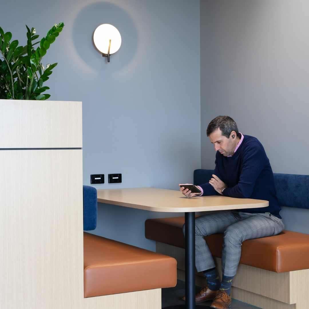 A man wearing a navy sweater and checkered pants is sitting in a booth at a modern, minimalist coffee shop. He is focused on his smartphone, perhaps checking the latest company growth updates from BUMA Australia. There is a potted plant on a partition next to the booth and a wall sconce providing soft lighting above him.