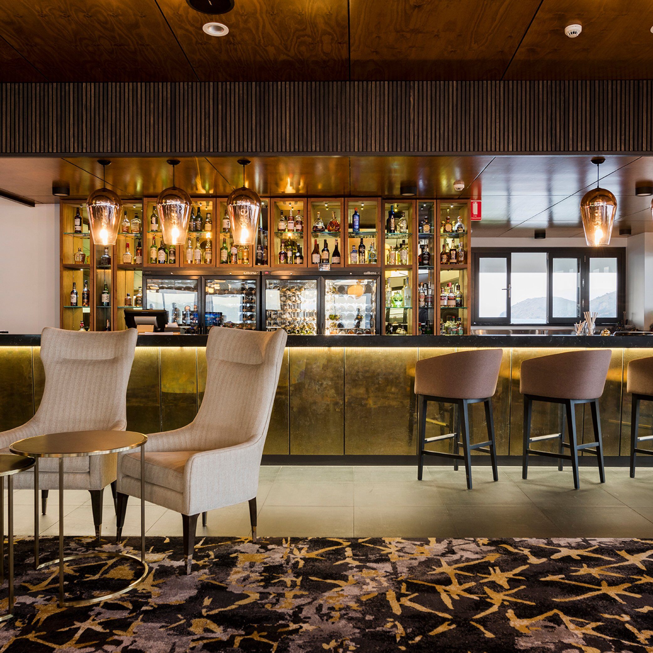 Stylish bar interior with a well-stocked backlit liquor display, pendant lighting, beige lounge chairs, and high stools along the bar counter.