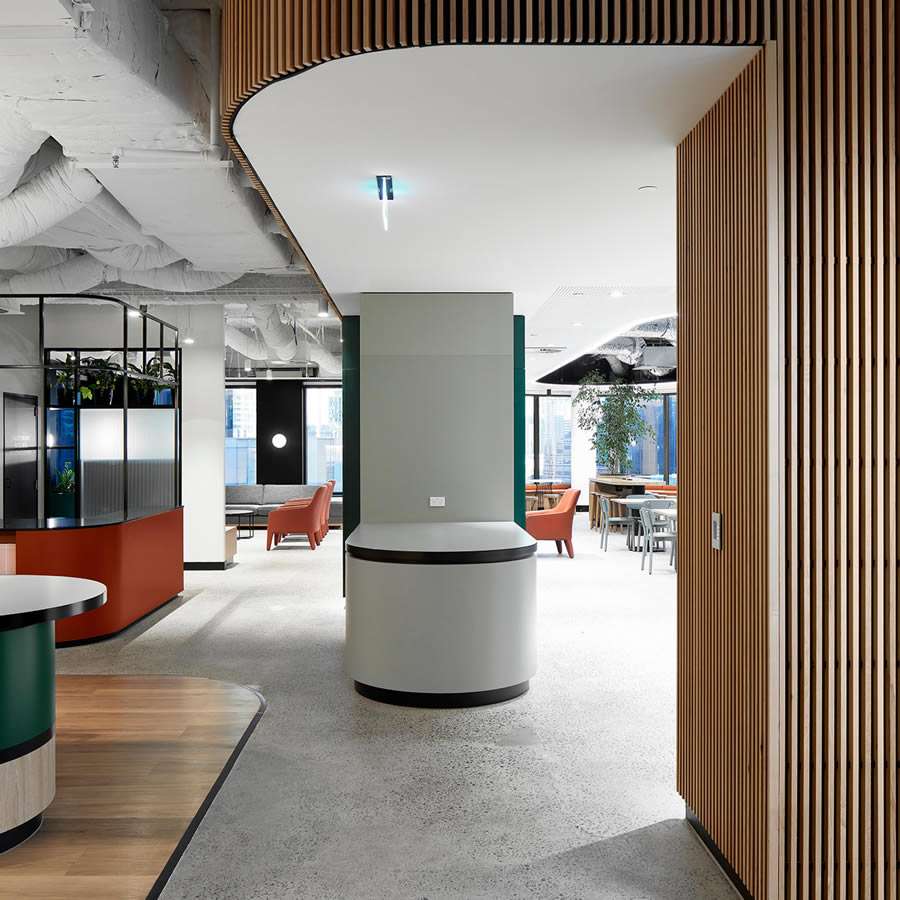 Modern office interior with an open floor plan featuring a mix of open workspaces and lounge areas, inspired by the shared neighbourhood concept. The design includes wooden slats, curved walls, and large windows allowing natural light. Furnishings are contemporary, with plants adding a touch of greenery.