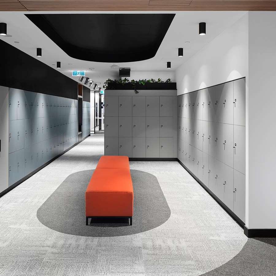 A modern hallway featuring rows of gray lockers on both sides, a long orange bench centered on a gray carpeted floor, and a black ceiling with recessed lights. The end of the hallway, designed with a shared neighbourhood concept in mind, has some greenery and a small exit sign visible above a doorway.