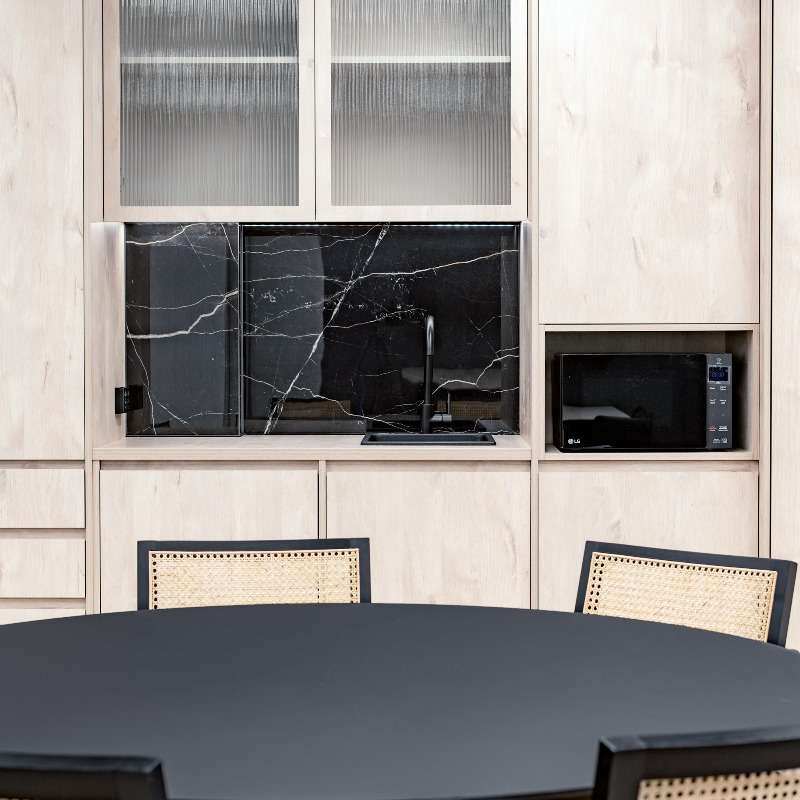 A modern kitchen in an Investa Sydney CBD property features light wooden cabinets and a sleek black marble backsplash. There is a black sink below the cabinets and a microwave on the right side. In the foreground, a round black table with four rattan-back chairs is visible, offering elegant comfort.