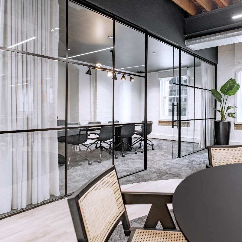 A modern Sydney CBD property featuring a glass-walled conference room with a long table and black chairs. The room has white curtains on one side, exposed wooden ceiling beams, and a large potted plant. Outside the conference room are additional chairs and a black round table, perfect for speculative suites.