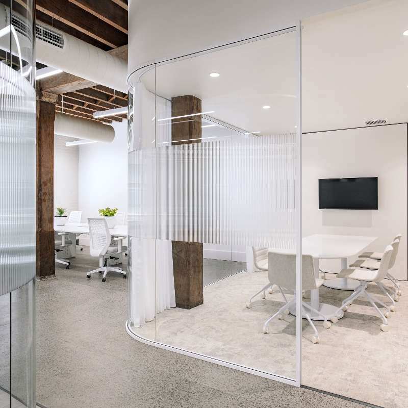 A modern, minimalist office space with white walls and concrete floors in an Investa Sydney CBD property. It features a glass-walled conference room with a wall-mounted TV and white furniture. Adjacent is an open workspace with white chairs, desks, and potted plants adding a touch of greenery.
