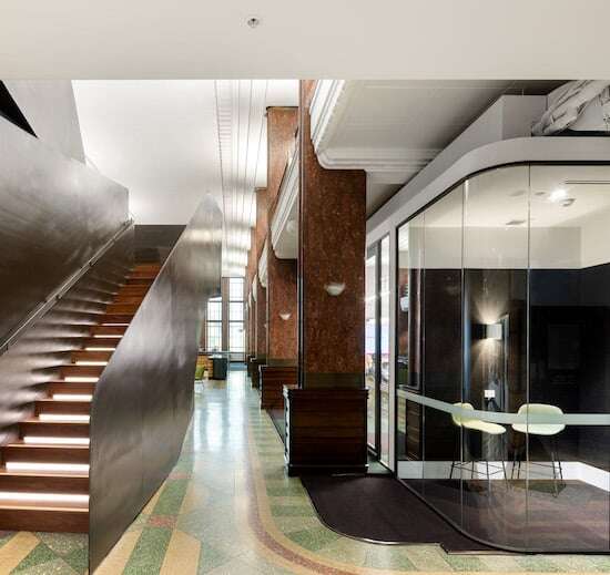Modern office interior featuring a sleek staircase with illuminated steps, a glass-enclosed meeting room perfect for collaboration with a small table and chairs, and a mix of contemporary and classic architectural elements including high ceilings and decorative columns—ideal for M&C Saatchi's hybrid workplace model.