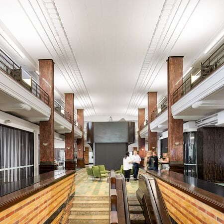 A spacious, well-lit interior features tall columns and a high ceiling. The room includes brick and glass accents, with green chairs arranged near the center. Several people are standing and walking in the background, providing a sense of scale to the space, embodying M&C Saatchi's innovative hybrid workplace culture.