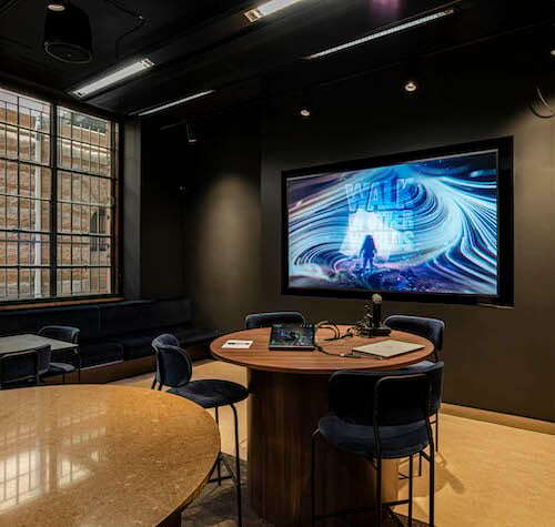 A modern, dark-toned meeting room at M&C Saatchi with a large screen displaying visuals on the wall. The round table in the center is surrounded by navy blue chairs, and there's a laptop, notebook, and pen on the table. Large windows on the left let in natural light, creating an ideal space for collaboration.