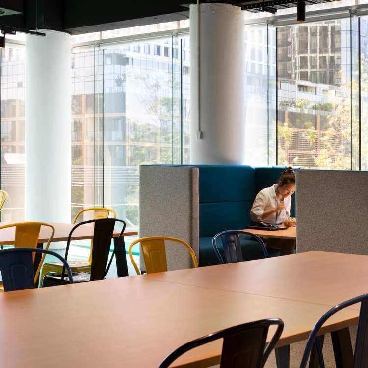 A spacious, well-lit office break area with large windows and modern furniture, ideal for fostering innovation. A person is sitting in a booth, working or reading at a table. Multiple chairs are arranged around large communal tables, and tall pillars add to the open and airy feel of the office space.