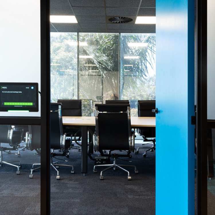 A modern conference room with a long table and multiple black leather chairs, viewed through a glass wall. There's a digital booking display on the left side of the glass wall. Large windows in the background reveal palm trees and natural light streaming in, reflecting an atmosphere of innovation.