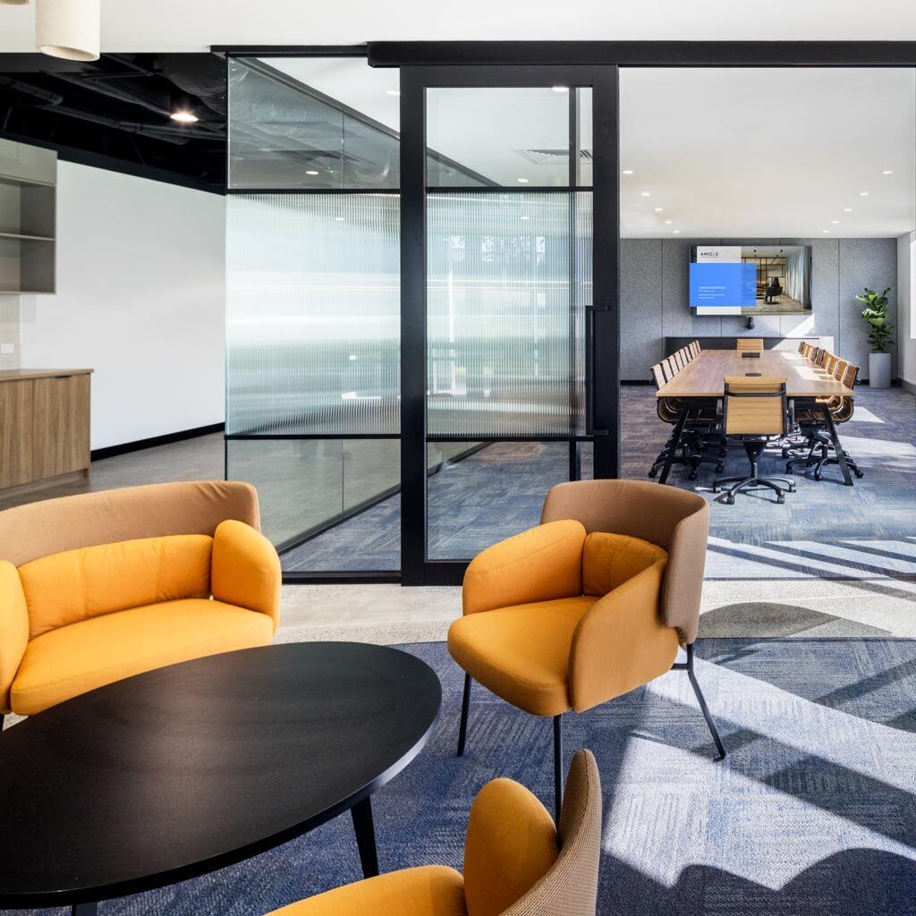Modern office space with a glass-walled conference room, featuring a long table, tan chairs, and mustard-yellow lounge chairs around a black coffee table in the foreground.