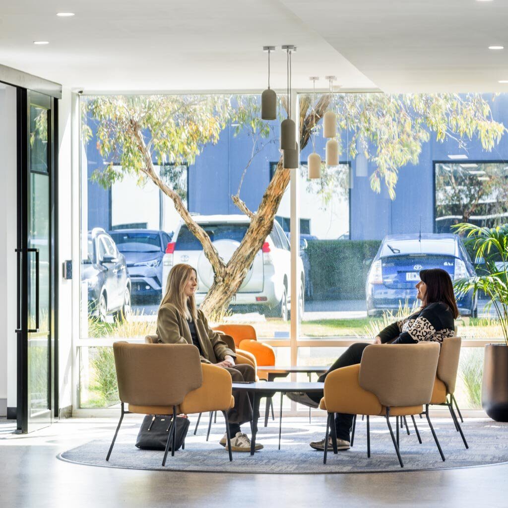 Two women sit and engage in conversation in the modern office lobby area of Amicus, a curated space with large windows allowing in natural light. They are seated in comfortable lounge chairs around a small coffee table, with cars and a tree visible outside.