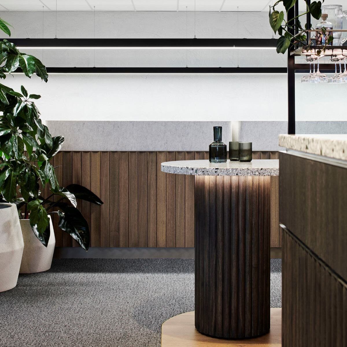 Modern interior space featuring a round table with a marble top, wooden base, and decorative bottles. Nearby, lush green plants in white pots and sleek wood paneling add a touch of nature. Overhead, contemporary lighting complements the neutral grey and brown tones, showcasing the FutureSpace aesthetic.