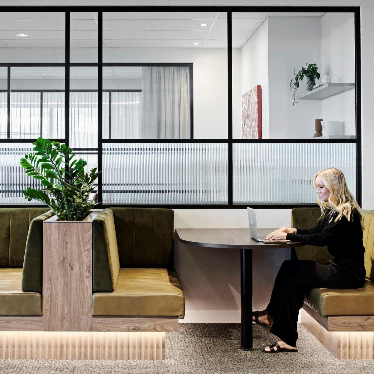 A woman sits at a corner booth in a modern FutureSpace office, working on a laptop. The booth has green upholstered seating and a large plant is placed on a divider. Black-framed glass panels separate the seating area from another office space in the background.