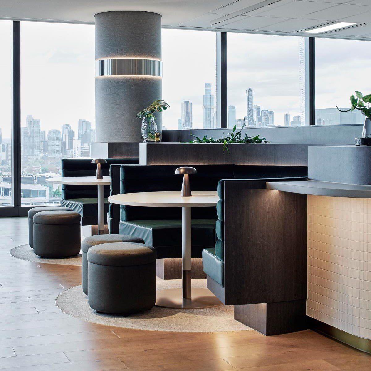 A modern office lounge area with round tables and green upholstered seating booths from Tradeflex, complemented by small poufs. Large windows offer a view of a city skyline, and plants add a touch of greenery. The design features clean lines and neutral tones.