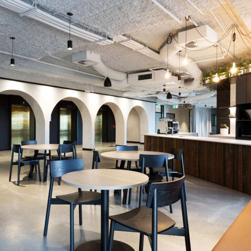 A modern cafe with minimalist decor featuring wooden tables, black chairs, and a bar counter with hanging lights. The ceiling has exposed pipes and ducts, while the wall has arches. With plants hanging above the counter and smart workplace technology implementation, the space exudes a relaxed, open ambiance.
